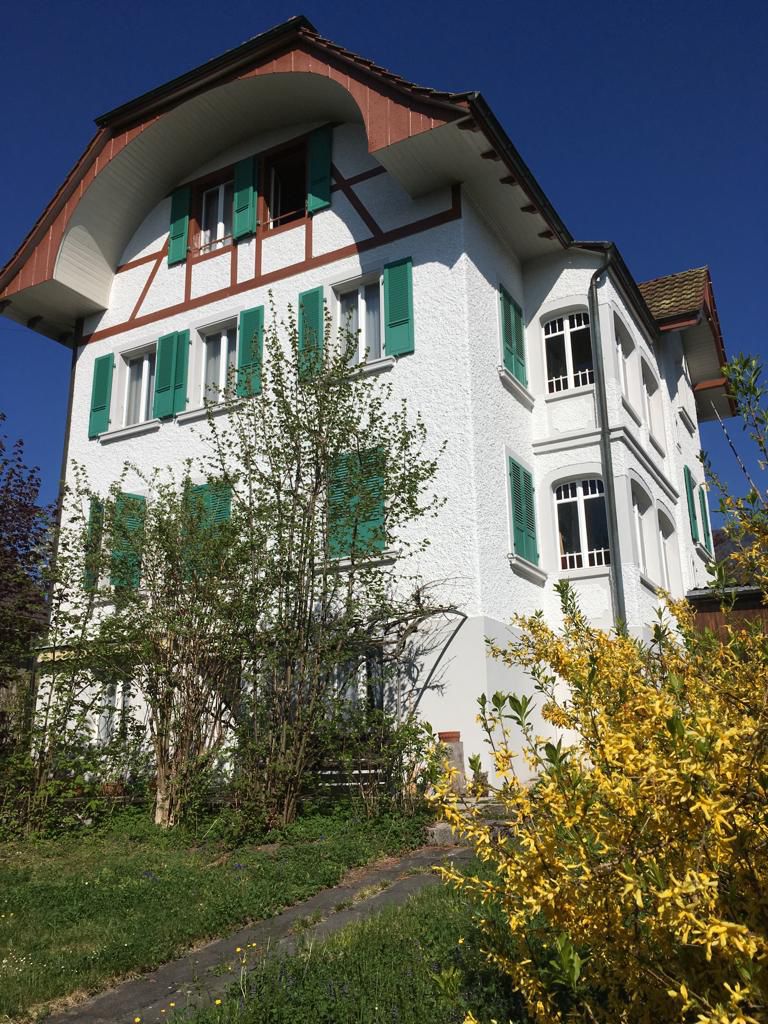 Two-story house with a traditional Swiss-style architecture, featuring a red-tiled roof, white walls, and green shutters. The house has a balcony or terrace on the upper level. The surrounding garden is lush with trees and shrubs, including a yellow flowe