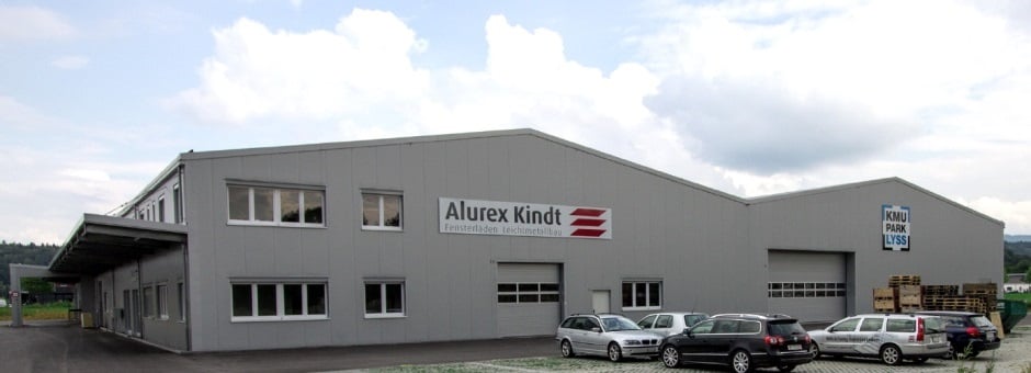 Large gray industrial building with the Alurex Kindt logo, multiple windows, and a parking lot in front of the building