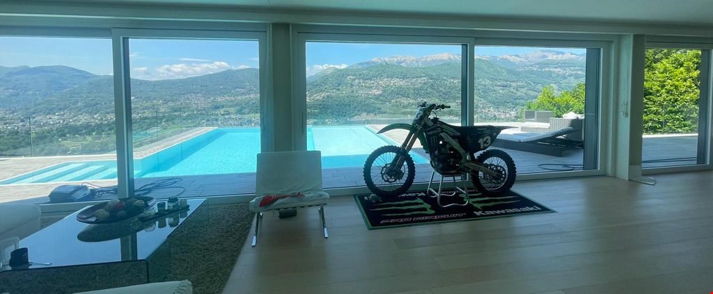 Large open-concept living room with floor-to-ceiling windows offering a panoramic view of the mountains and a swimming pool outside. The room features a modern design with a dirt bike displayed on a platform in the center.