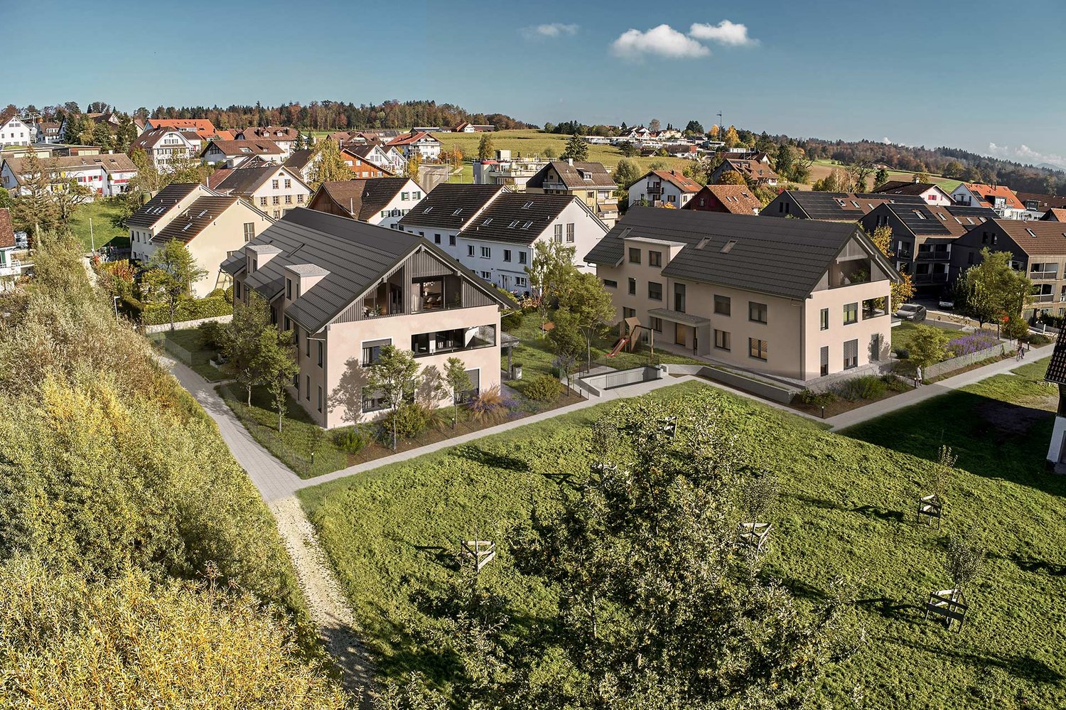 The image shows a scenic residential area in a hilly, forested region. The buildings are a mix of multi-story apartment buildings and smaller, single-family homes with red tile roofs. The buildings have balconies and are surrounded by lush greenery, inclu