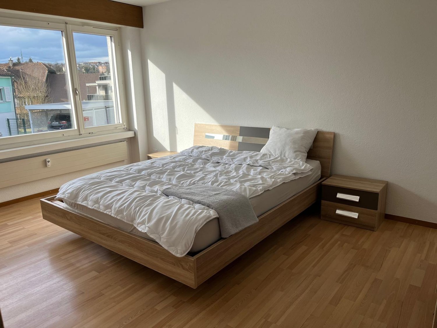 Spacious bedroom with hardwood flooring, large windows providing natural light, a wooden bed frame with a gray comforter, and two nightstands with drawers.