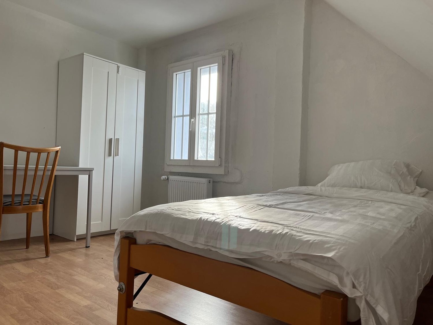The image shows a bedroom with a double bed, a white wardrobe, and a wooden chair. The room has a window that provides natural light, and the walls are painted in a light, neutral color.