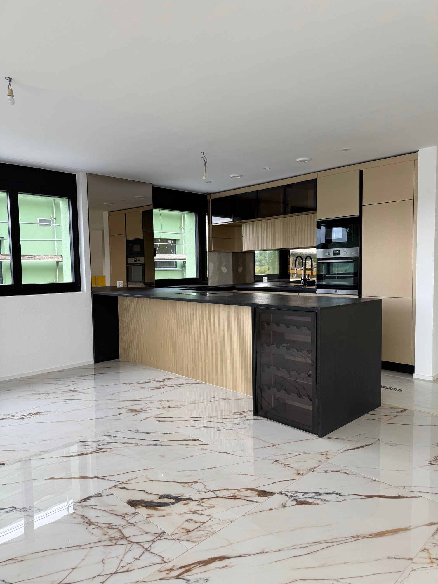 Modern kitchen with sleek black and beige cabinets, marble-patterned tile flooring, built-in appliances including a wine cooler, and large windows providing natural light.