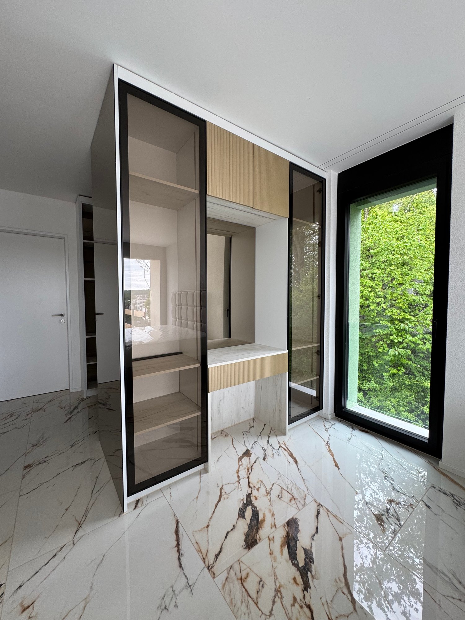 Spacious bedroom with large windows providing a view of the greenery outside. The room features built-in wardrobes with a mix of glass and wood panels, creating a modern and sleek design. The floor is covered in high-quality marble tiles, adding a touch o