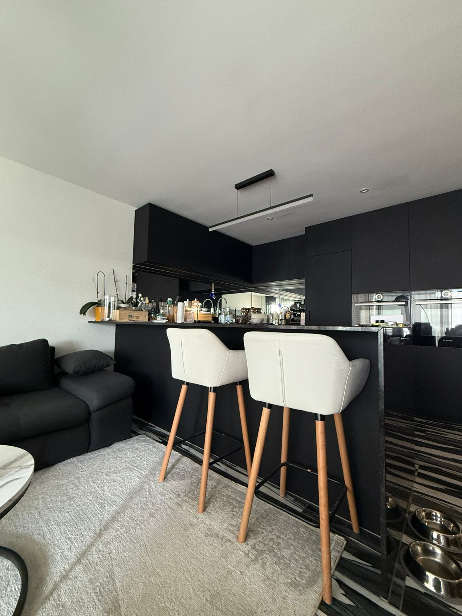 open kitchen with a marble bar, two white leather stools, a marble countertop, black cabinets, grey carpet, black sofa, and a grey coffee table
