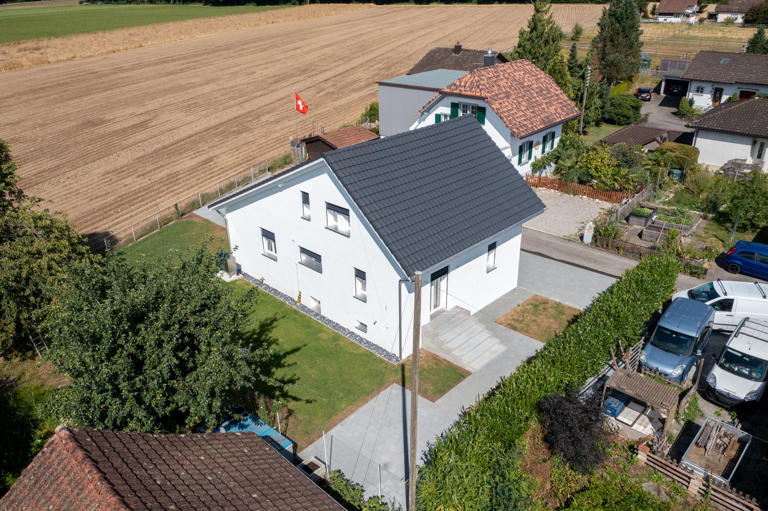 Two-story white house with a red tile roof, surrounded by a garden and trees. The house has a garage and parking space visible. The property is located in a rural area with a large open field in the background.