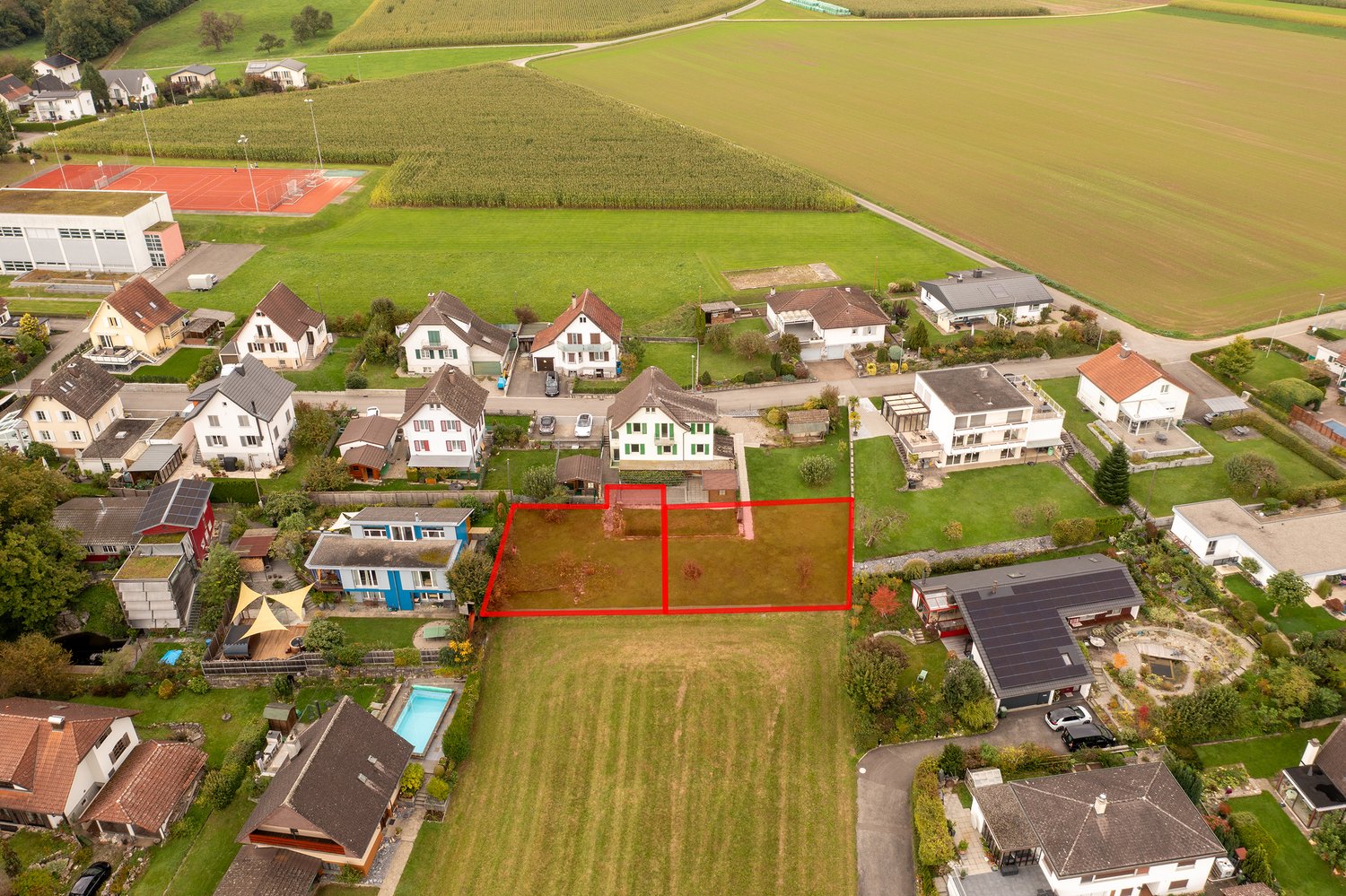 Houses in a residential area, with fields and a tennis court in the background. A swimming pool is visible in the bottom left corner.