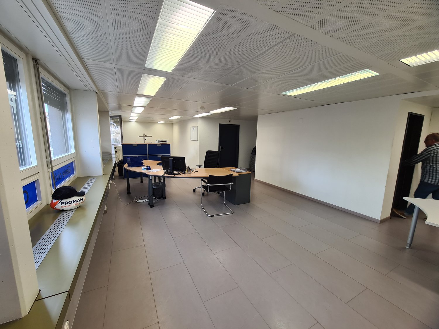 The image shows a modern, open-plan office space with a tiled floor, suspended ceiling with recessed lighting, and several desks with computers and office chairs. The space appears to be well-lit and organized, with a Proman sign visible in the corner.
