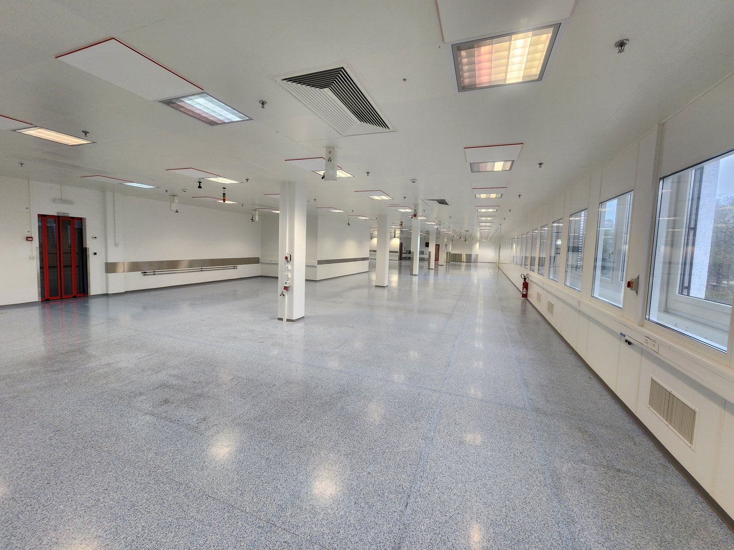 Long empty hall, white walls and ceiling, industrial style, multiple columns, windows on one side, fire extinguisher.
