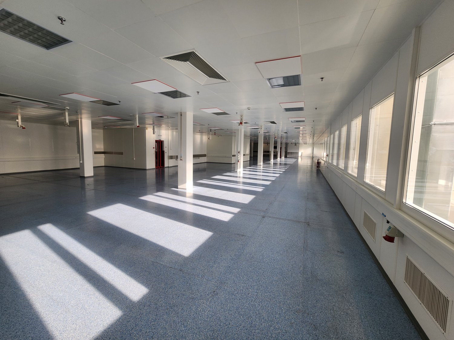 spacious room, white walls, high ceilings, gray flooring, large windows on one side, multiple red fire extinguishers