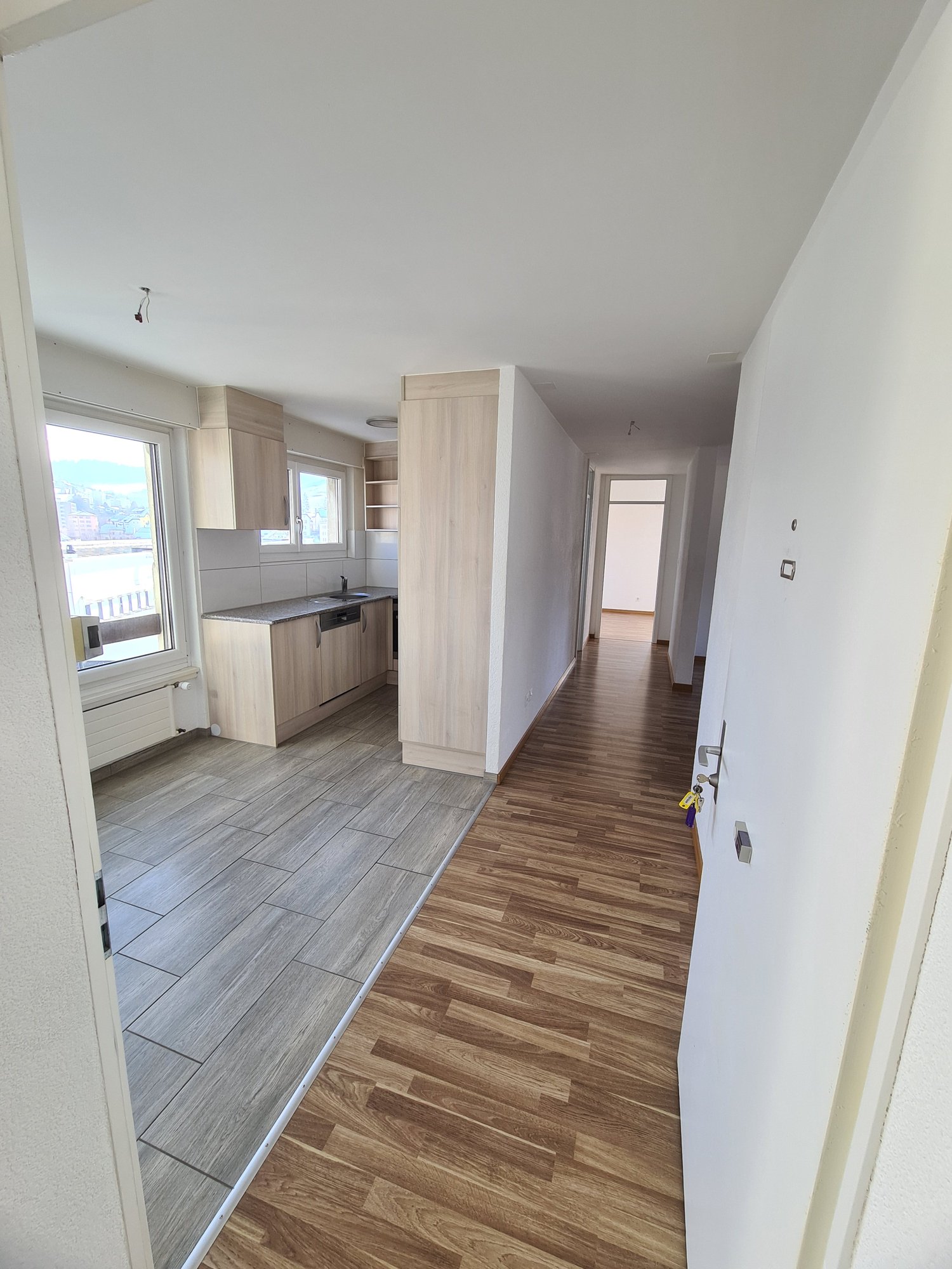Empty hallway with wooden floor, white walls, kitchen with sink, cabinets, and dishwasher