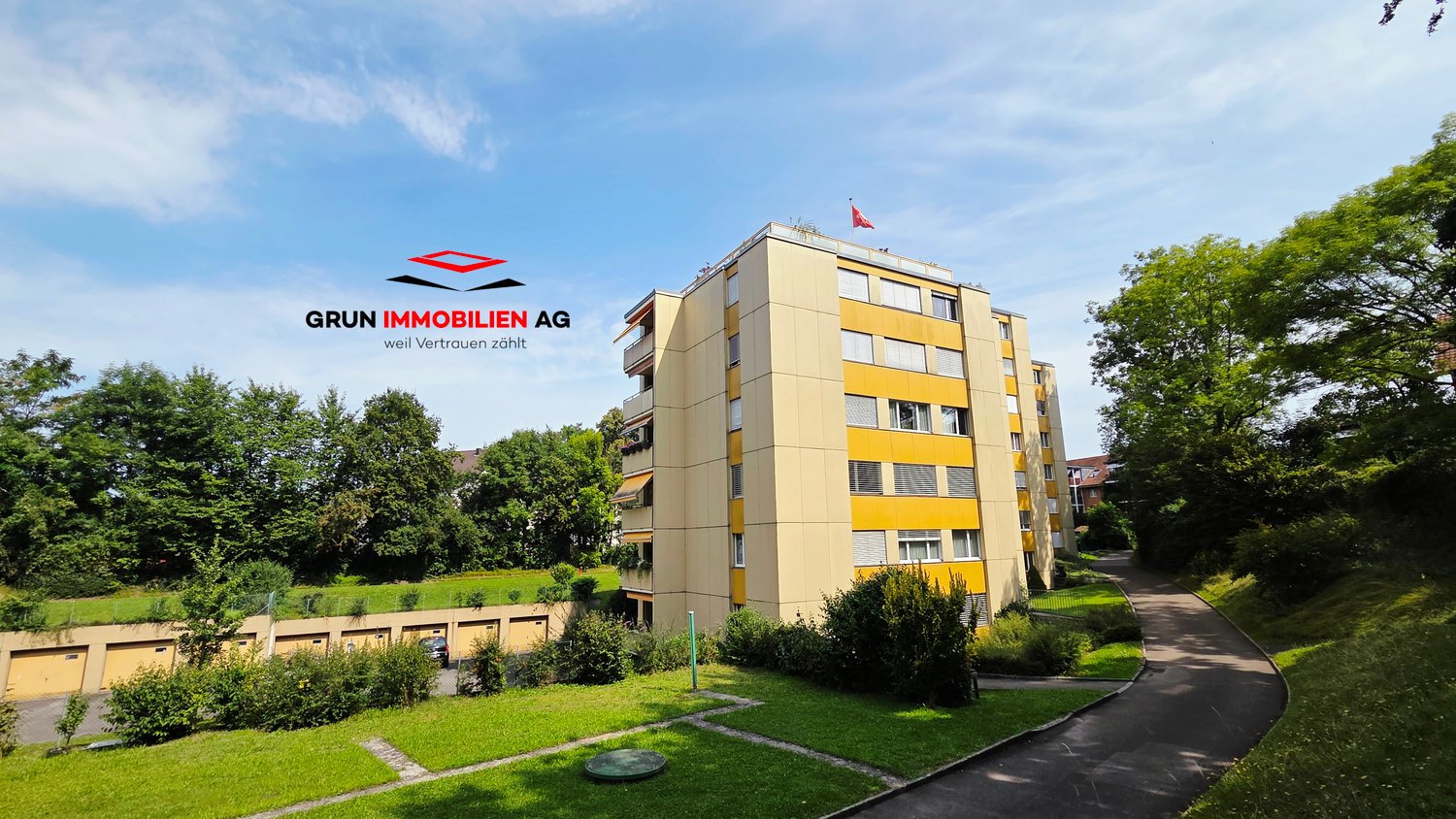 High-rise apartment building, yellow exterior, multiple floors, garage doors visible, a red flag on top, driveway leading to garage