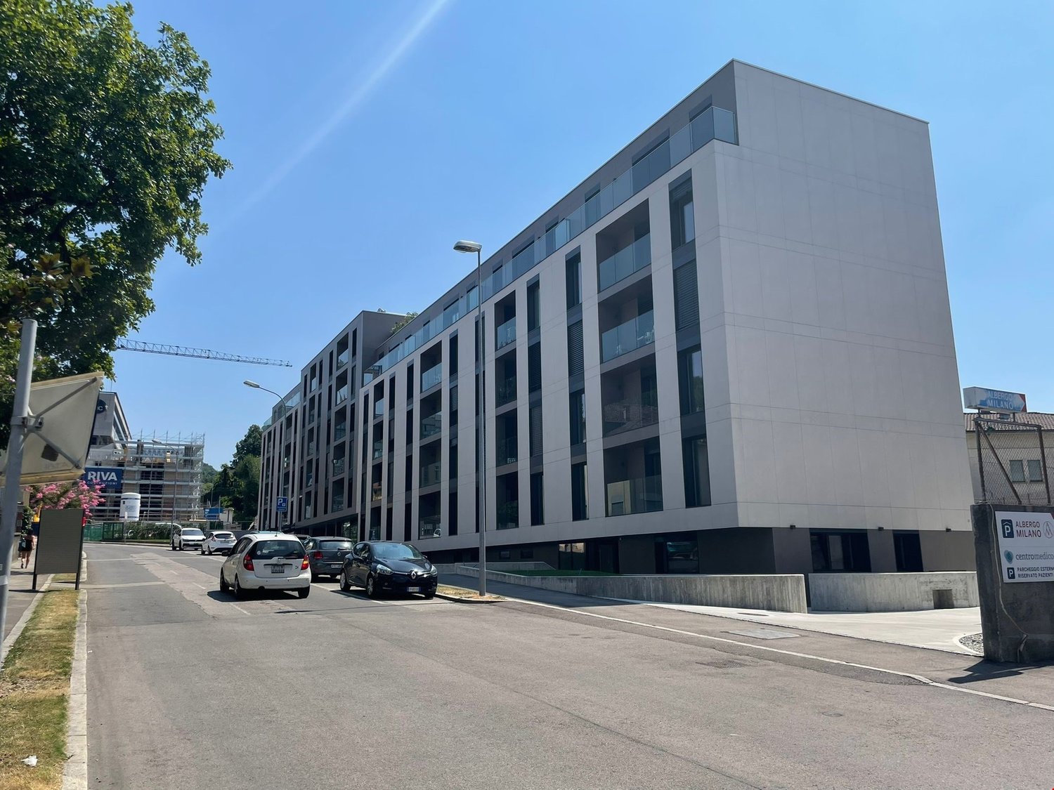 A modern multi-story commercial or residential building with a gray exterior and large windows. There is a parking area in front of the building with several cars parked.