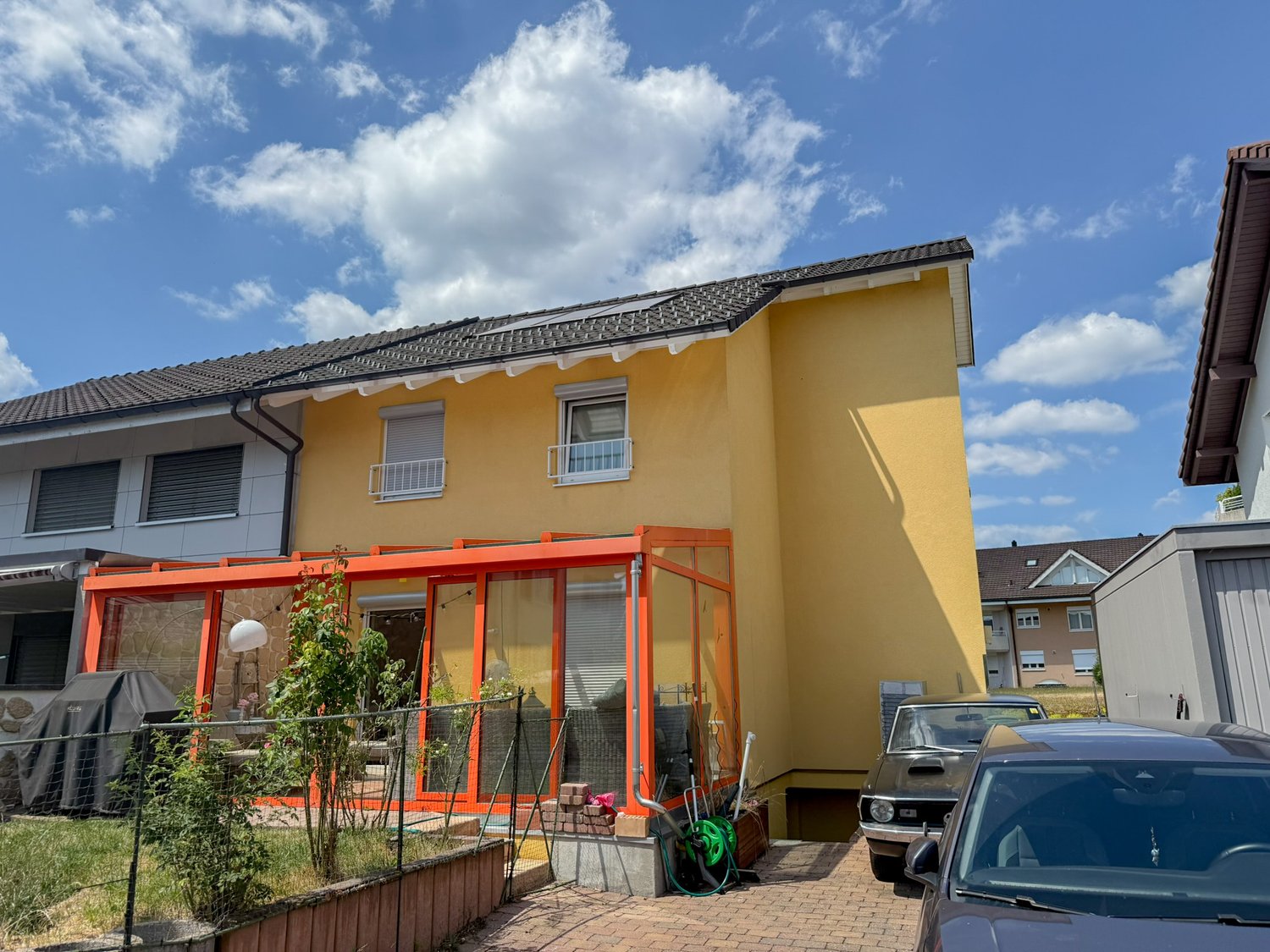 2-story yellow house with a red tiled roof, balcony, and a paved driveway with a car parked in front.