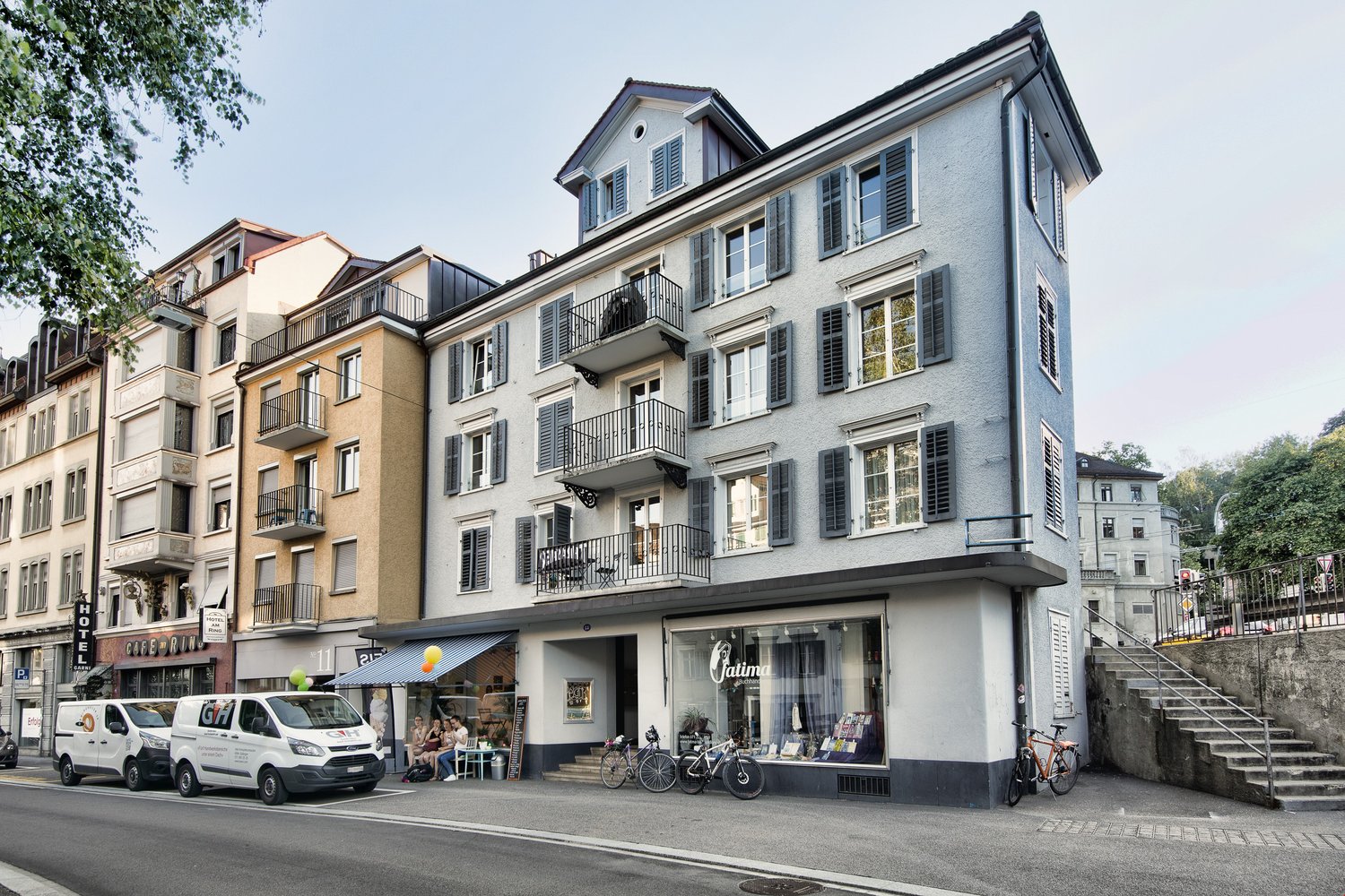 Apartment building, gray exterior, multiple balconies, storefront with awning, bicycles parked outside, stairs leading to upper floors, multiple vans parked on the street.
