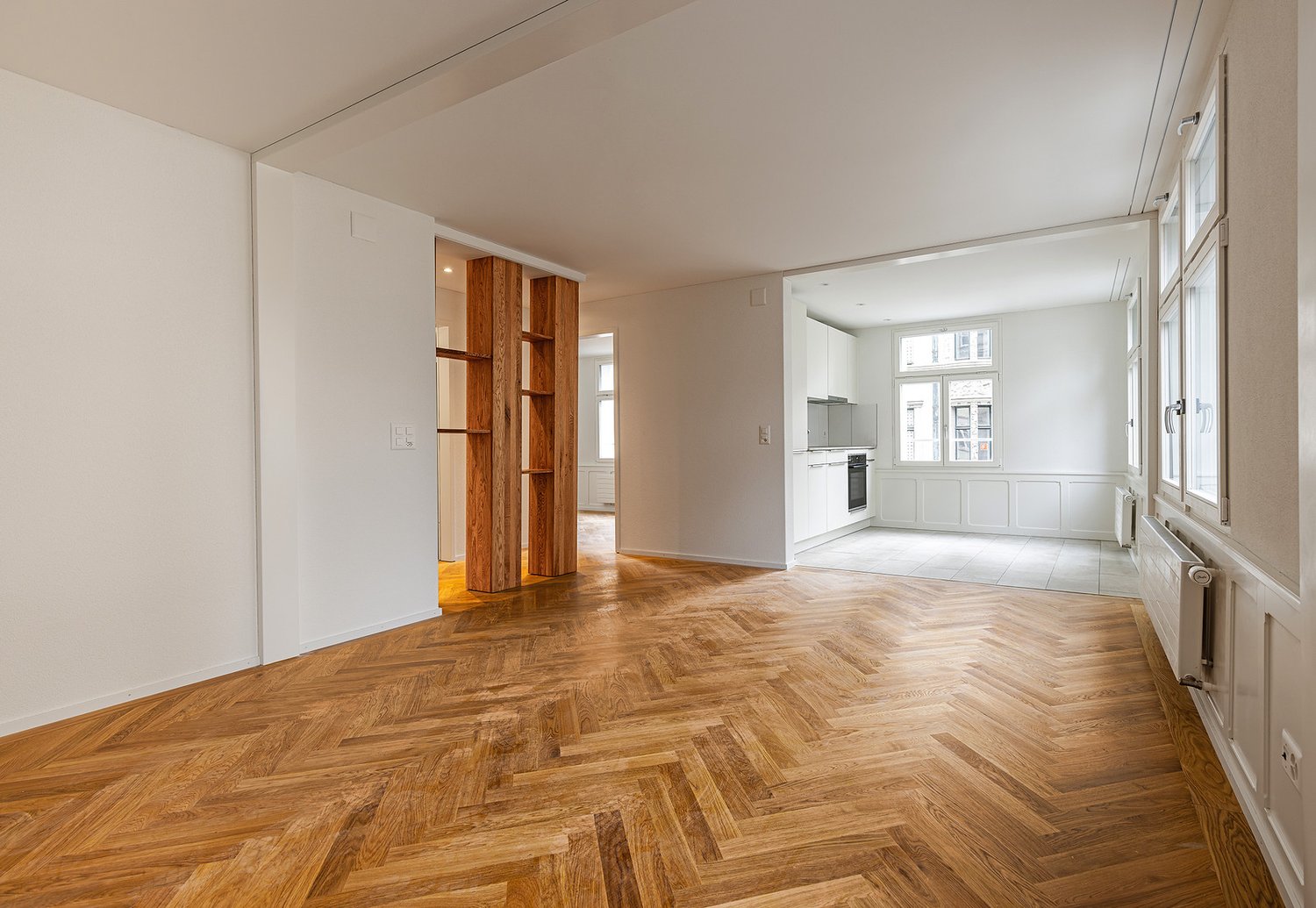 large room with wooden parquet flooring, white walls, wooden shelves, white cabinetry, radiators, and glass windows