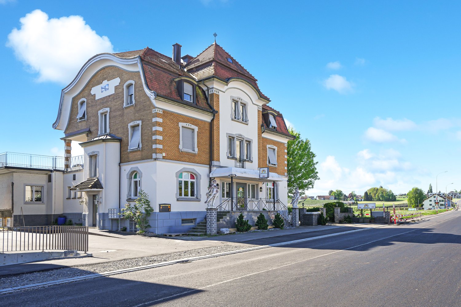 2 story house, brown roof, brick walls, windows with shutters, front garden, metal fence, stairs to the entrance, sign on the wall