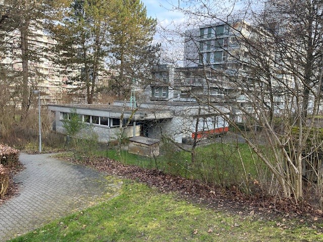 A building with many windows in the background, concrete floor, surrounded by many trees and a pathway