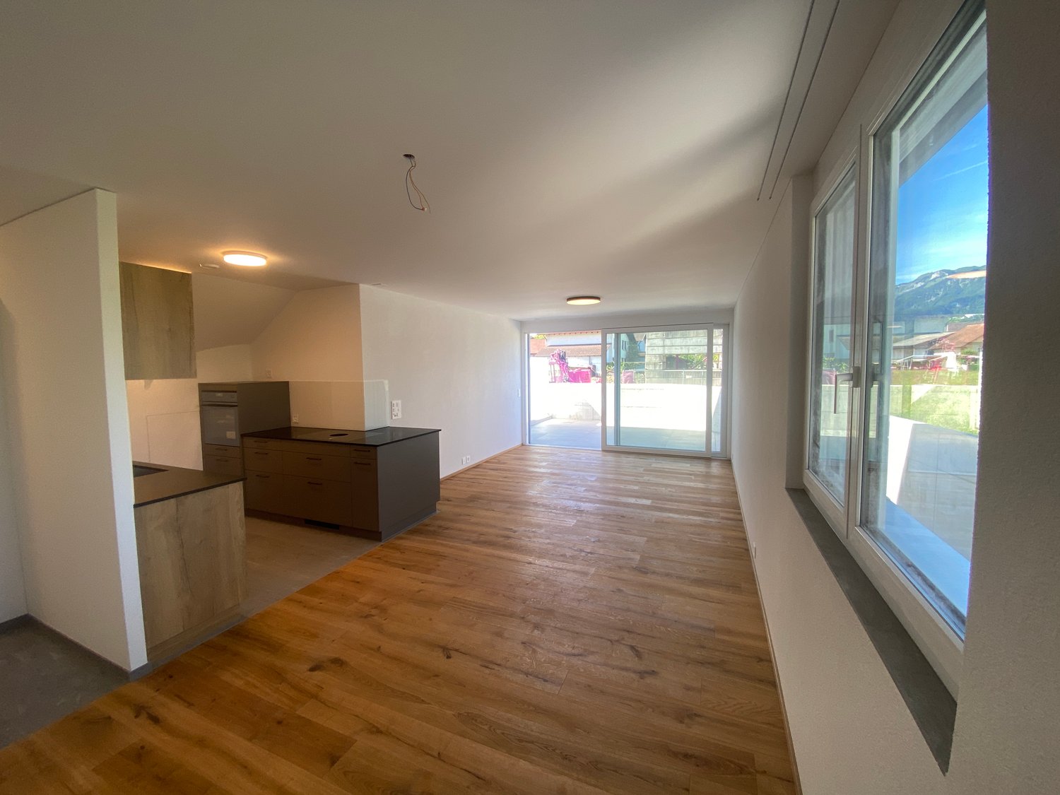 Open-plan living area with hardwood flooring, white walls, large windows providing natural light, and a sliding glass door leading to a balcony or terrace.
