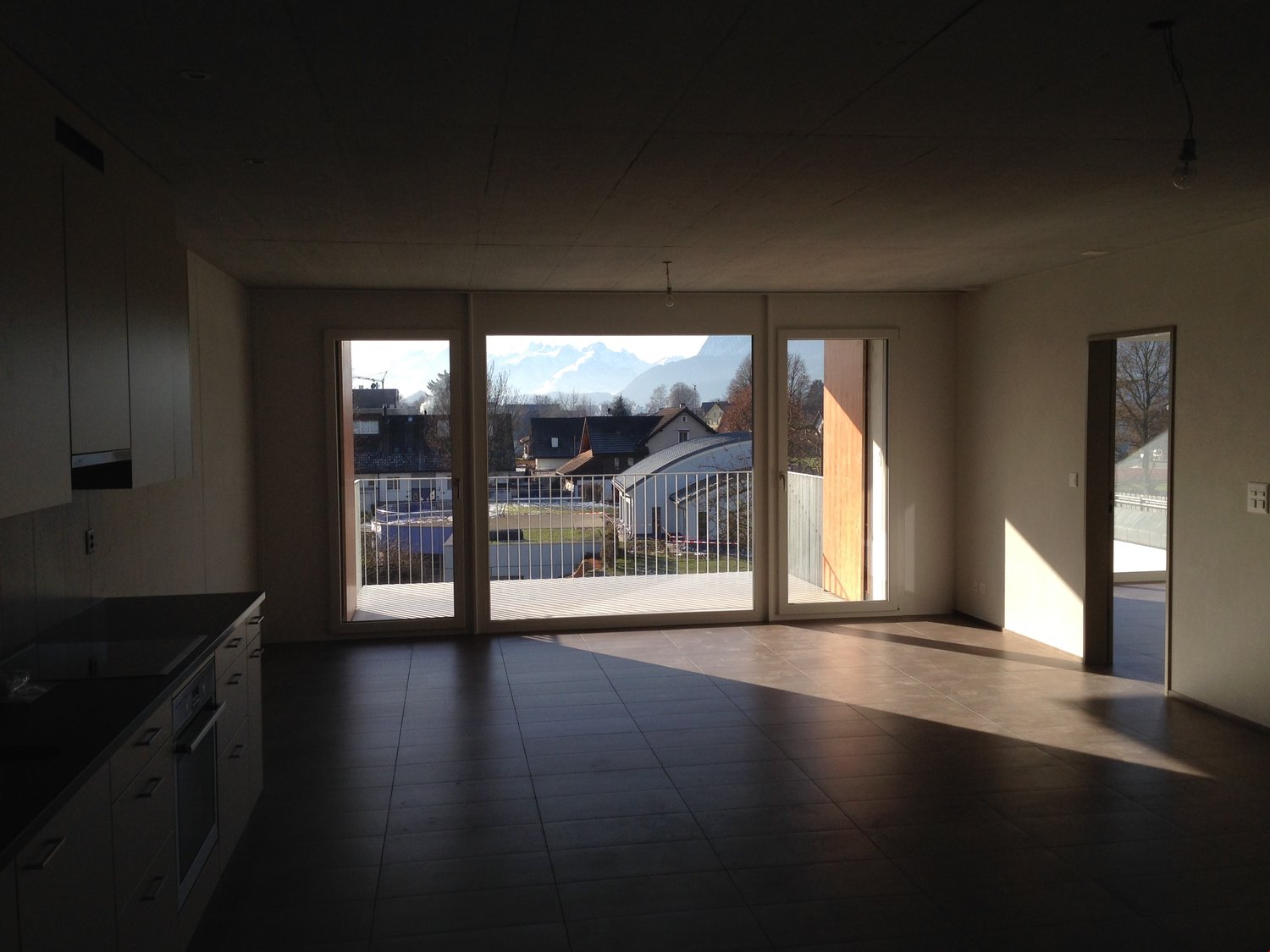 Spacious living room with large windows overlooking a balcony or terrace, dark tile flooring, and a view of the surrounding buildings and landscape outside.