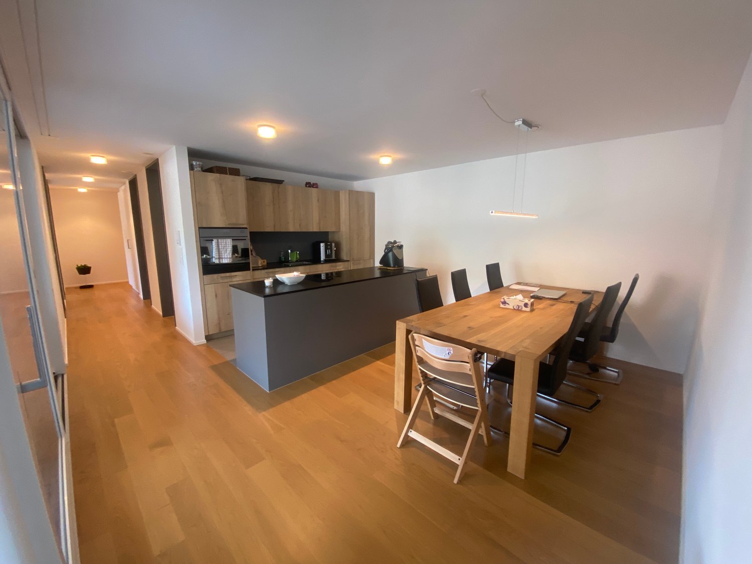 Open-concept kitchen and dining area with wooden cabinets, black countertops, and a wooden dining table with chairs. The kitchen has a dishwasher and other appliances. The space has hardwood flooring and recessed lighting.