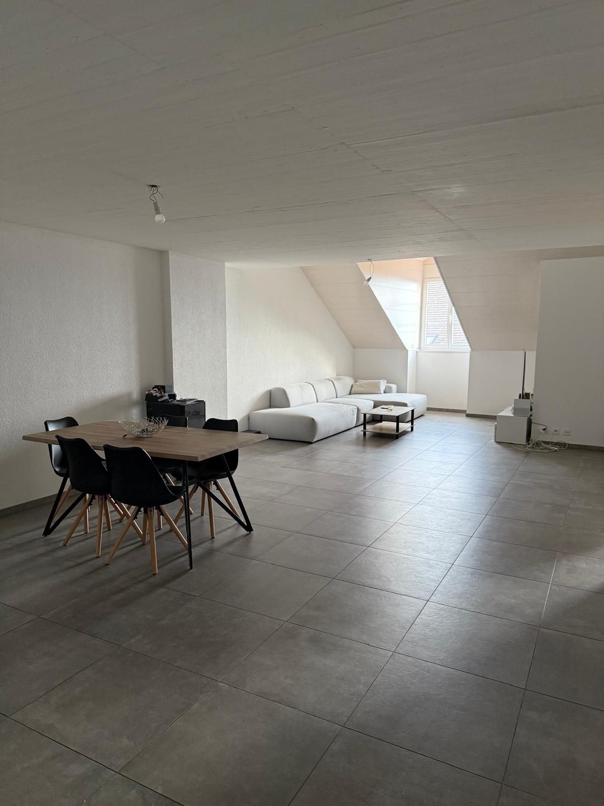 open living room, gray tiled flooring, wooden dining table and black chairs, white sofa with a coffee table, white walls, a light fixture, and a small window in the ceiling