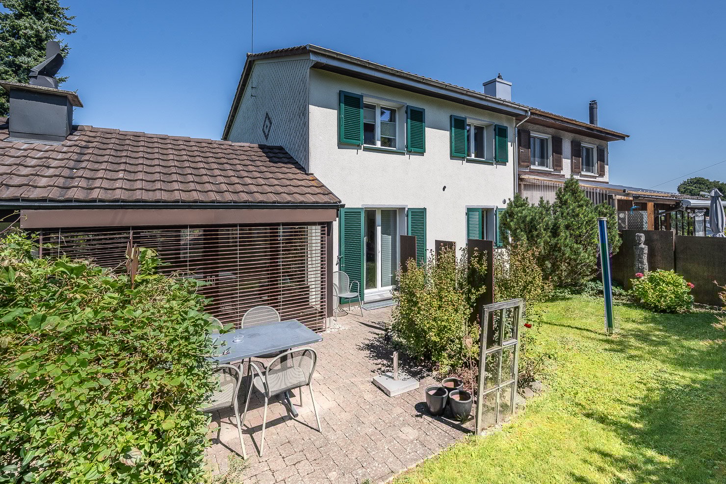 2 story house, brown tiled roof, white walls, raised ground floor, patio, greenery, chairs, table, small bushes, window