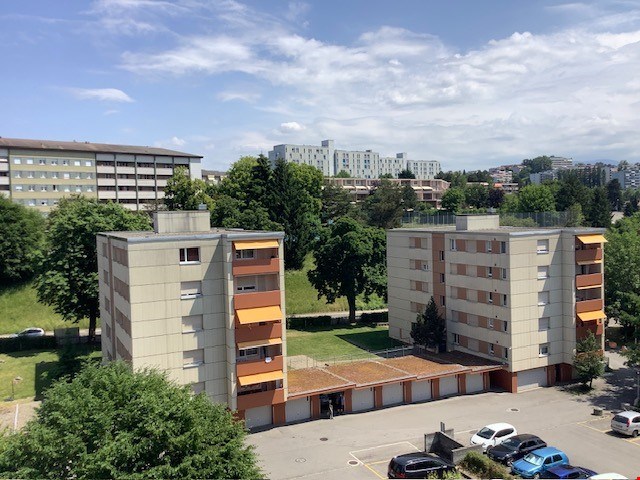 The image shows several multi-story residential apartment buildings with balconies and a parking lot in the foreground. The buildings appear to be in an urban setting, with some taller buildings visible in the background.