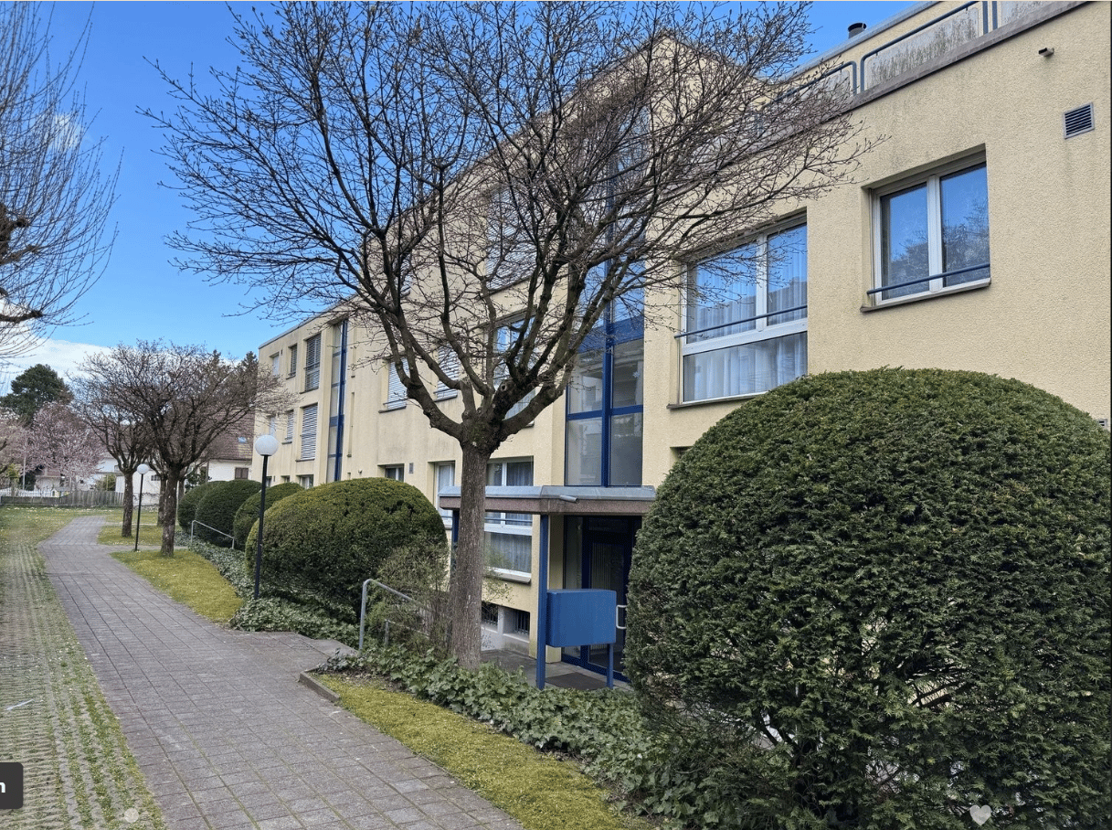 Multi-story apartment building with beige exterior, large windows, and surrounded by trees and landscaping. There is a paved walkway in front of the building.