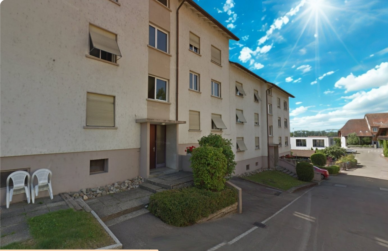 3-story apartment building with beige exterior, multiple windows, and a small garden area with shrubs in the front. The building appears to be located in a residential area with a paved parking area in front.