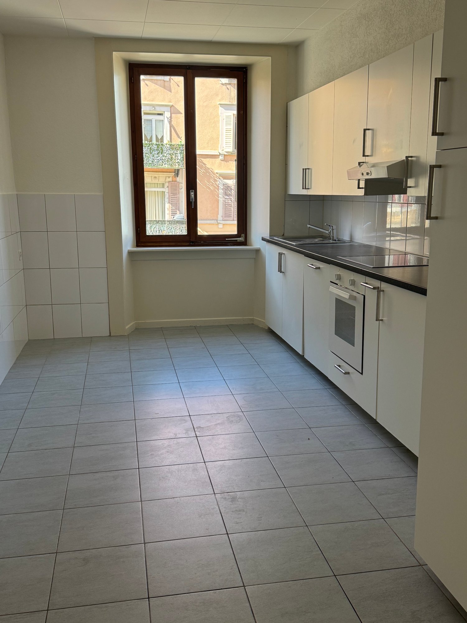 An empty kitchen with tiled floor, white cabinets, a window, a sink, and an oven