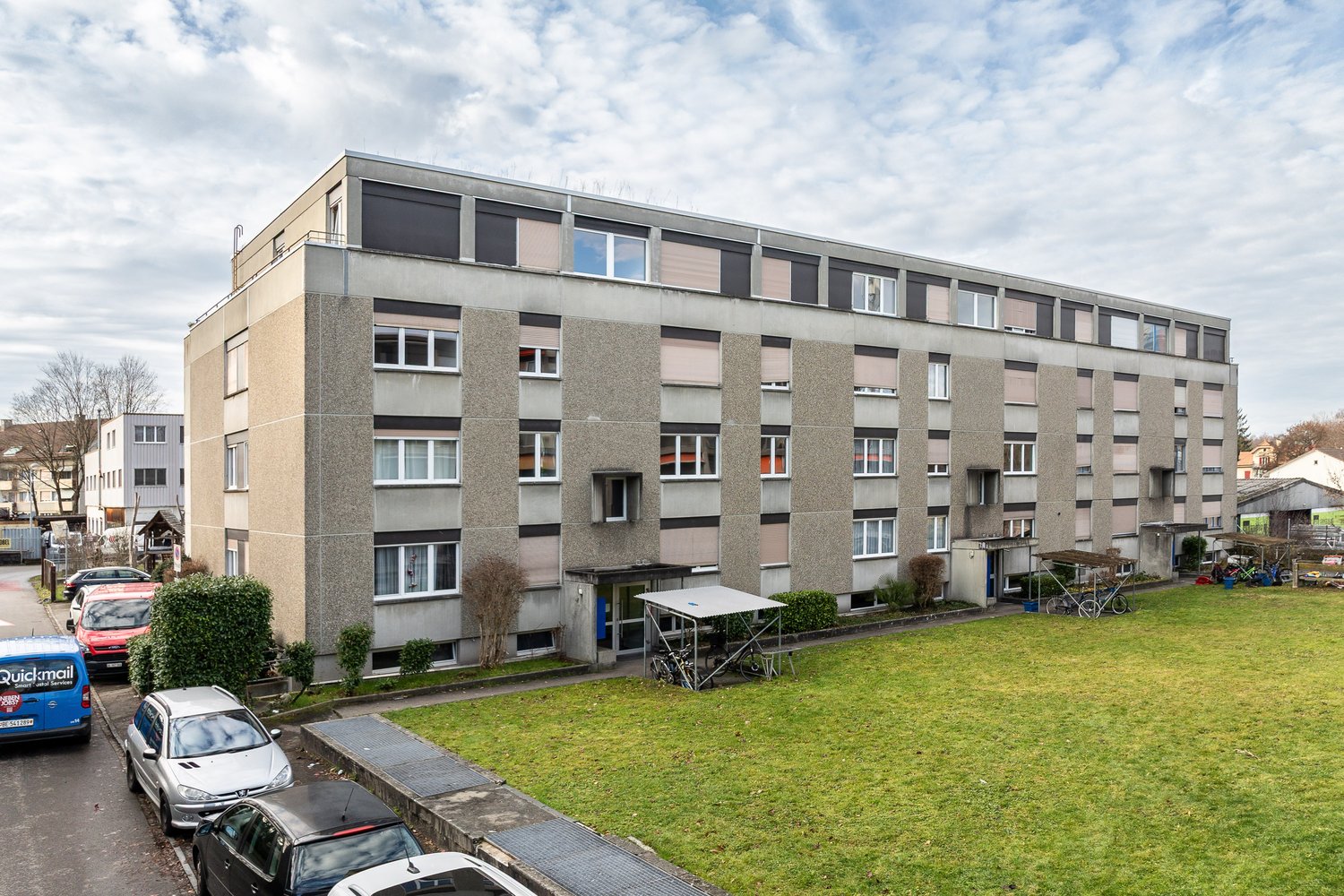 4 story apartment building, grey exterior, multiple windows, bicycles parked in front, small garden area