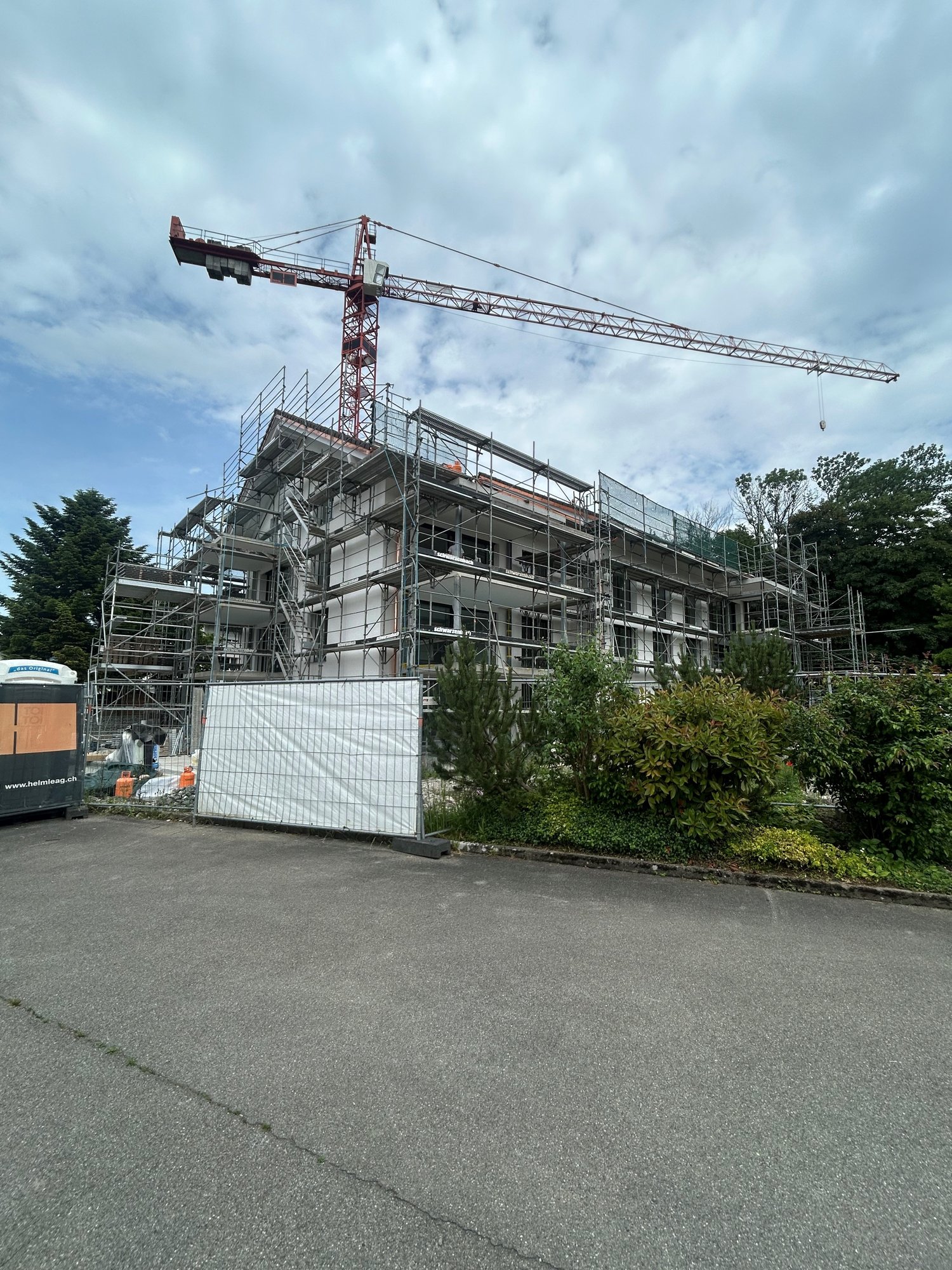 The image shows a construction site with a large crane and a partially built multi-story building. The building is surrounded by scaffolding and there is a fenced-off area in the foreground, likely for vehicle access or storage.