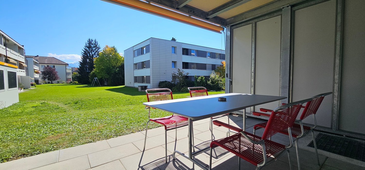 Outdoor dining table and chairs, lawn in front, modern apartment building behind, glass doors to balcony, clear skies, tree on lawn