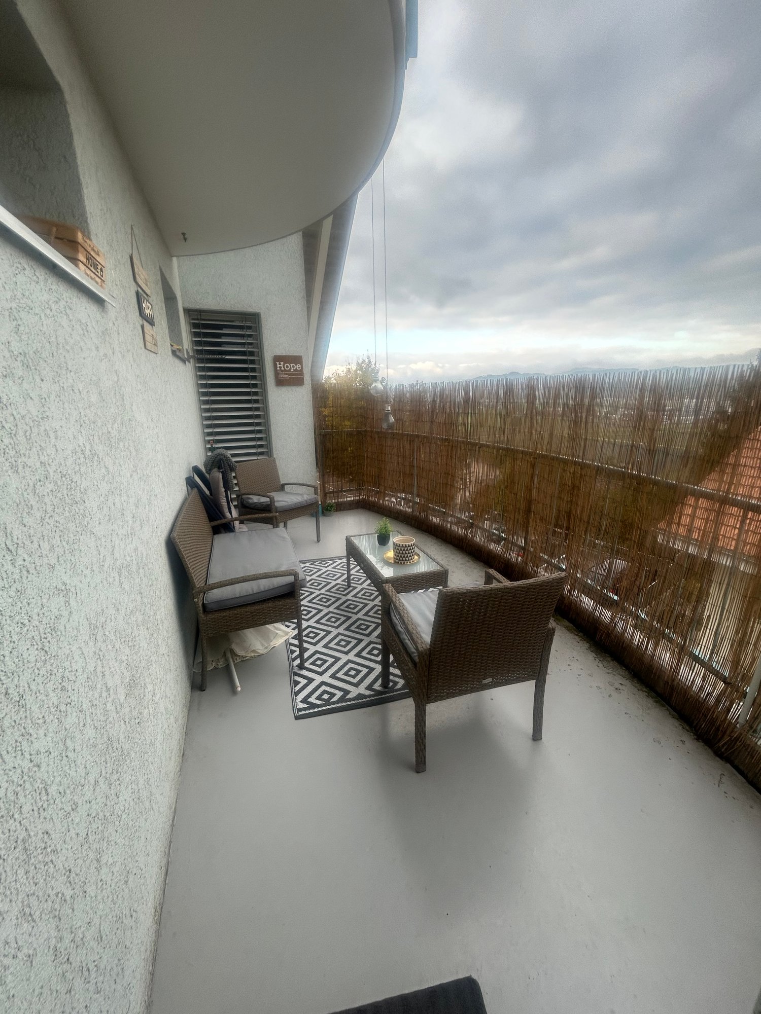 Balcony with outdoor furniture, black and white floor mat, glass table, plant in pot, gray walls, metal railings, cloudy sky