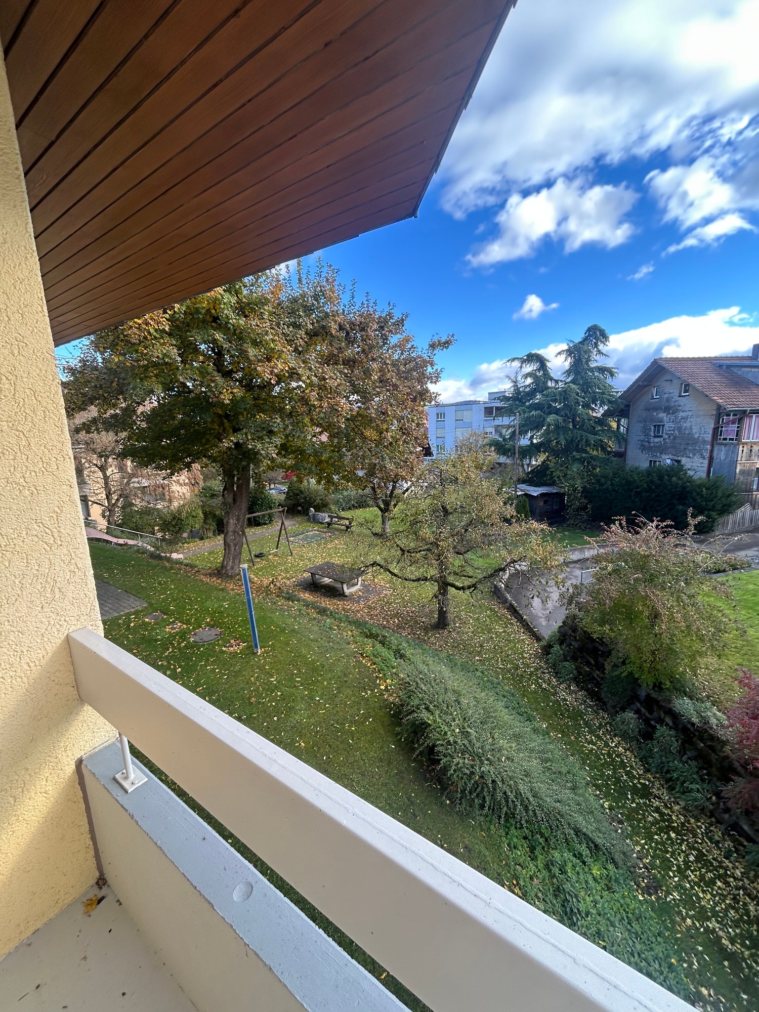 Balcony, overlooking a grassy area with trees and a path, clear blue sky, buildings in the background