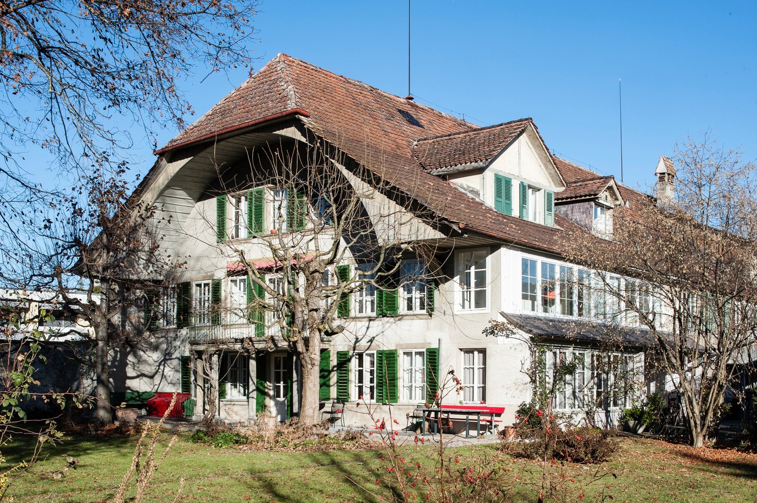 Two story house, red roof, green shutters, balcony on second floor, chimney, garden with benches