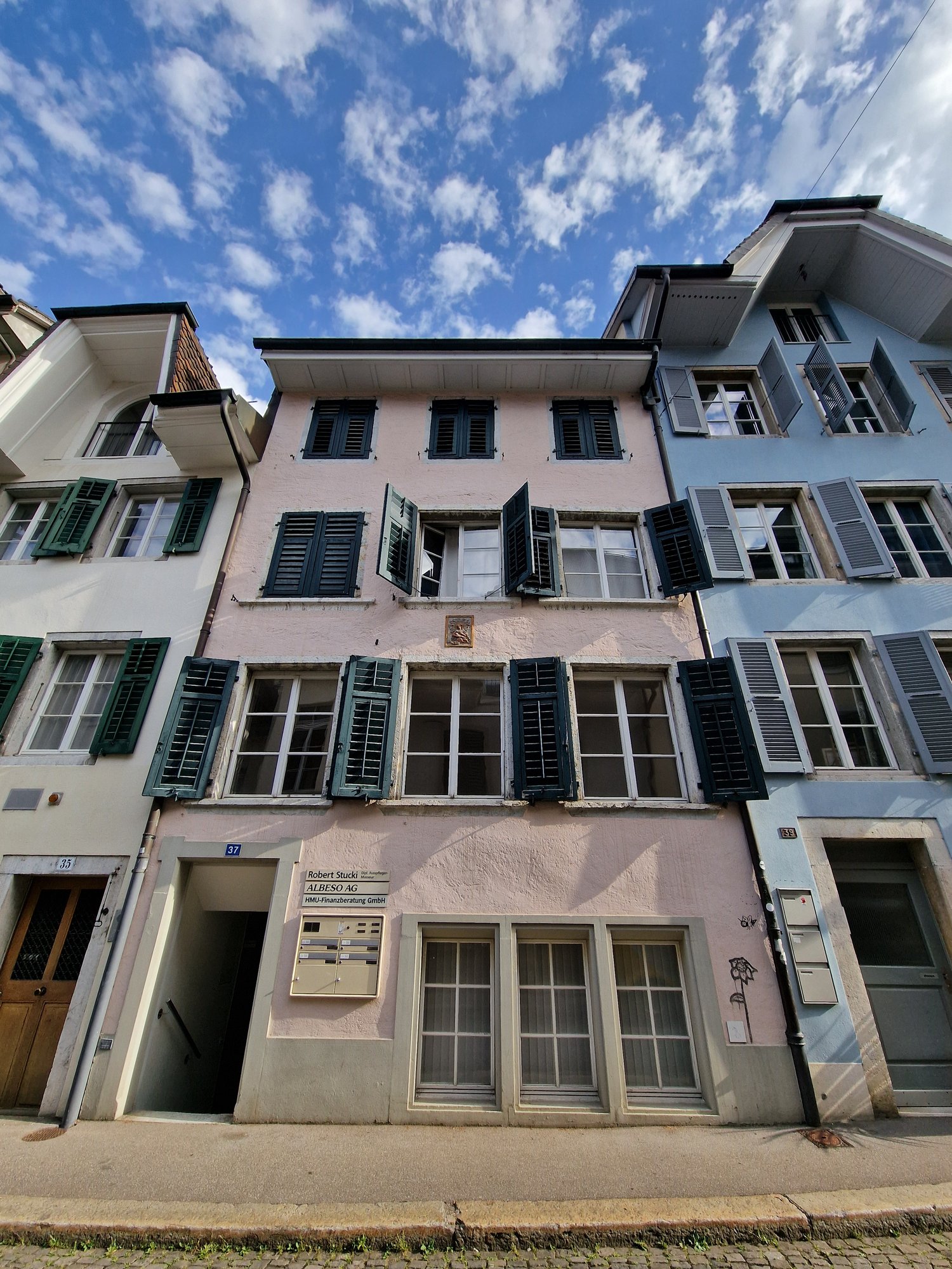 A multi-story building with a traditional European architectural style, featuring white walls, black shutters, and large windows. The building appears to be a commercial or office space, with a sign for 'Robert Stucki' on the ground floor.