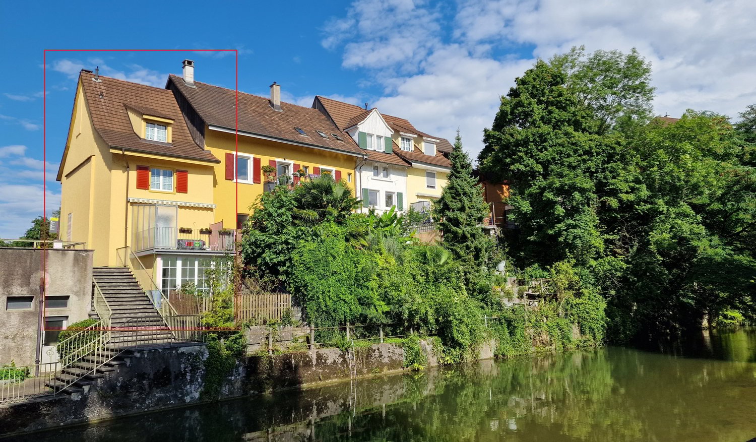 two-story house, yellow exterior, brown roof, red shutters, garden with stairs leading to river