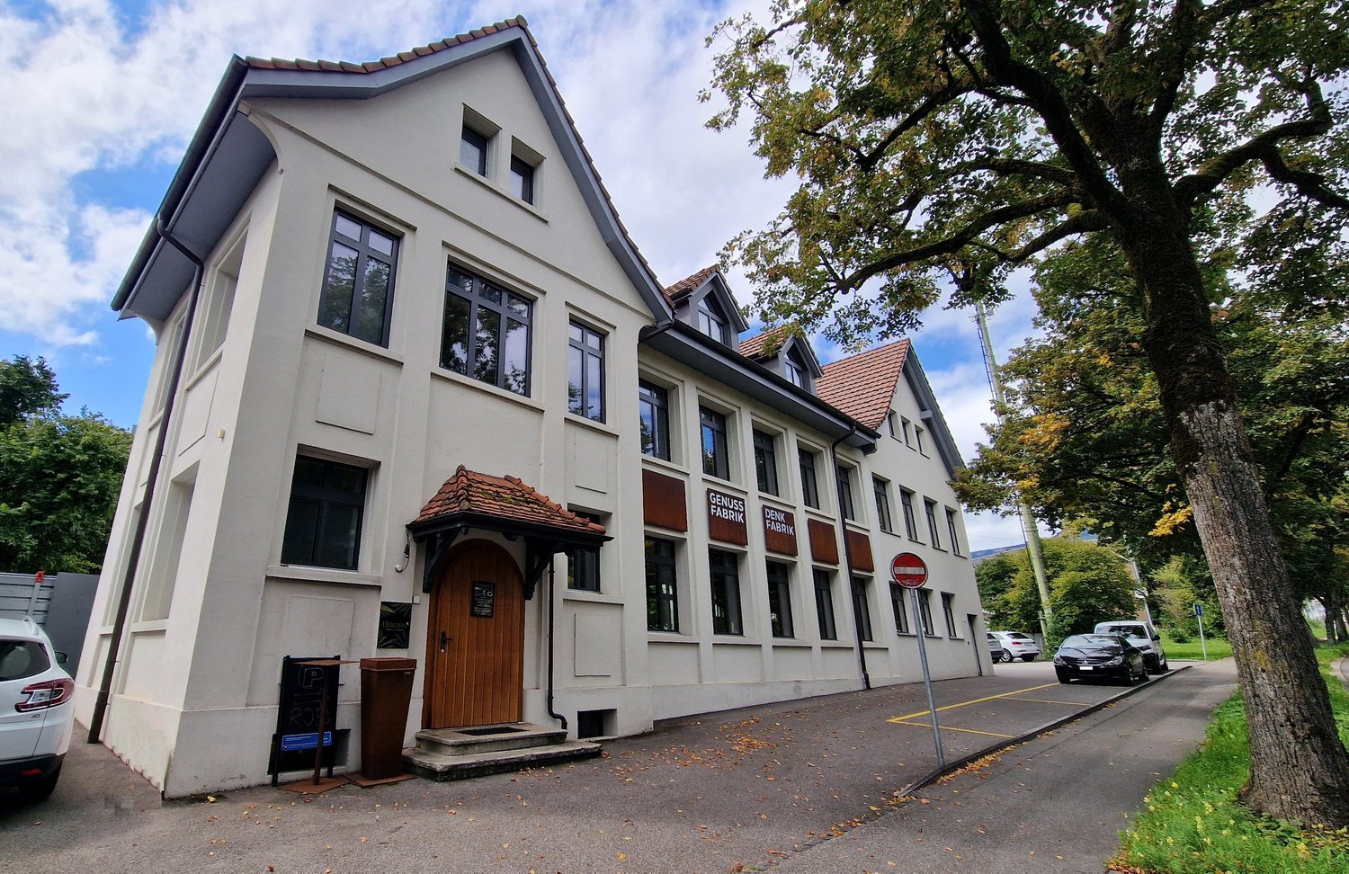 White house with brown roof, multiple windows, entrance door, trees, cars parked on the street