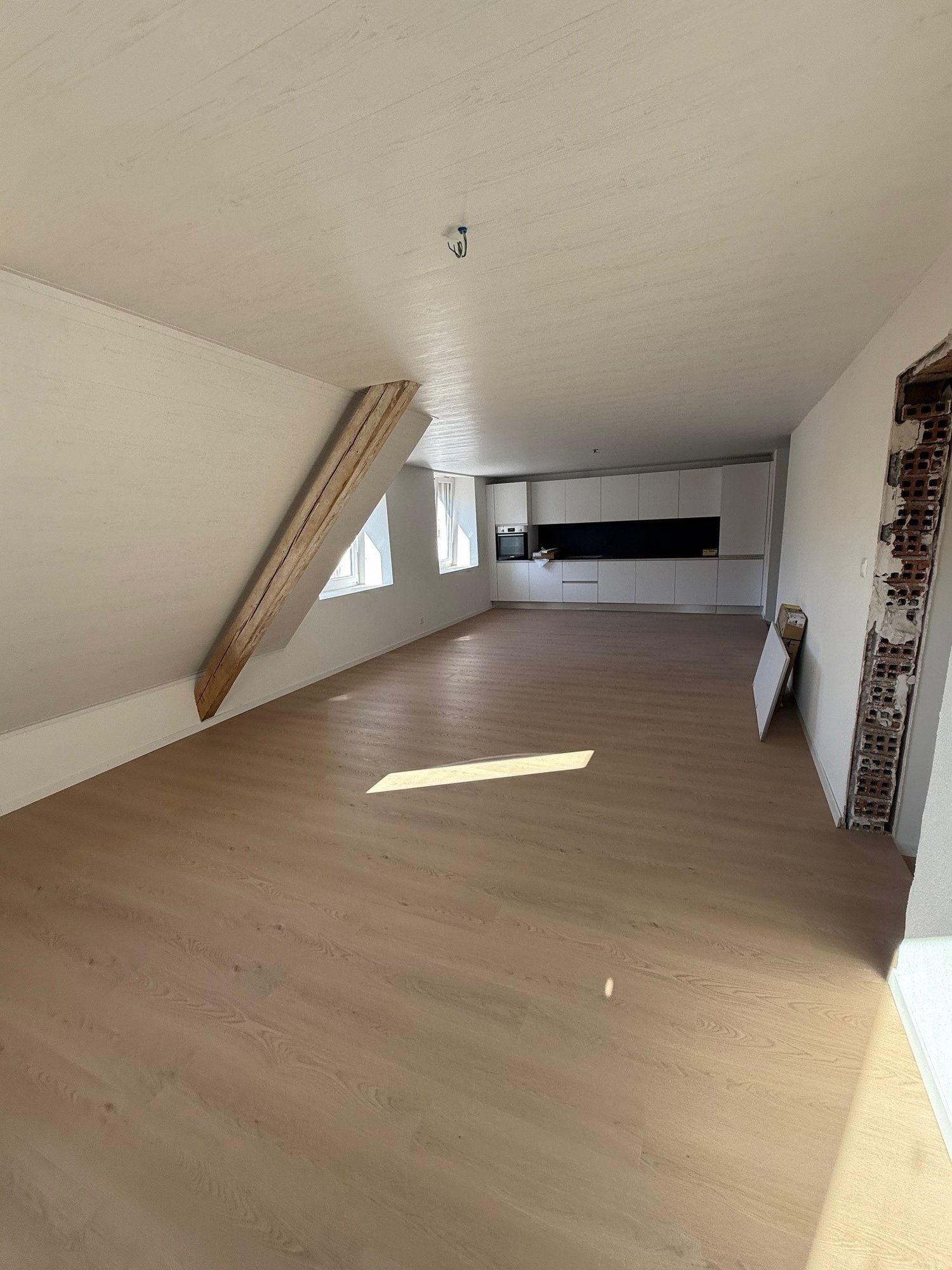 Open-concept living space with white walls, hardwood floors, and a modern kitchen with white cabinets and black countertops. The room has large windows that allow natural light to enter the space.