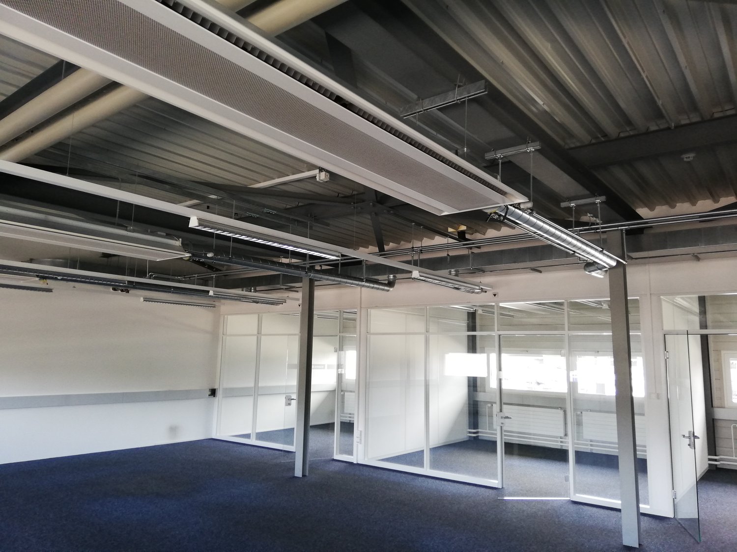Large open space with office walls and glass doors, metal ceiling with overhead pipes and lighting