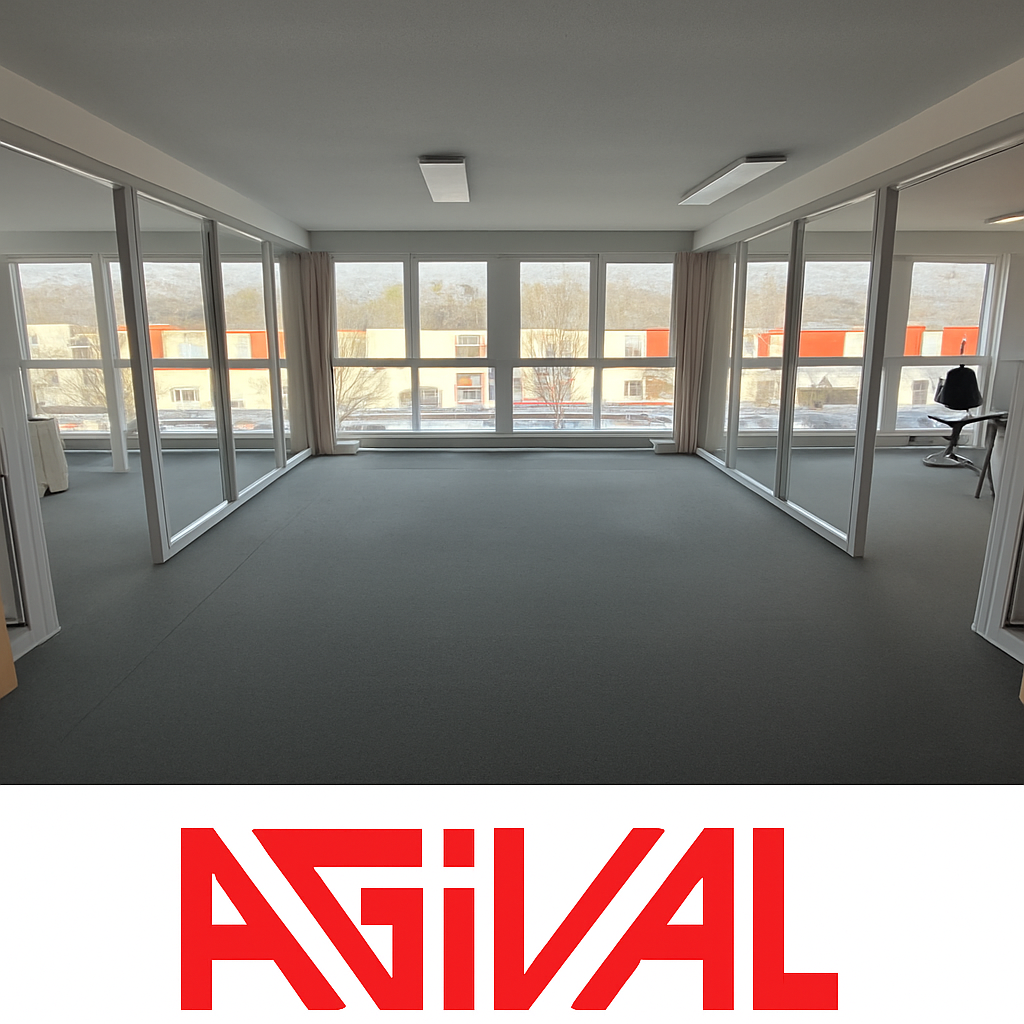 Large open office space with floor-to-ceiling windows providing a view of the surrounding buildings. The room has a gray carpet floor and a desk chair visible in the corner.