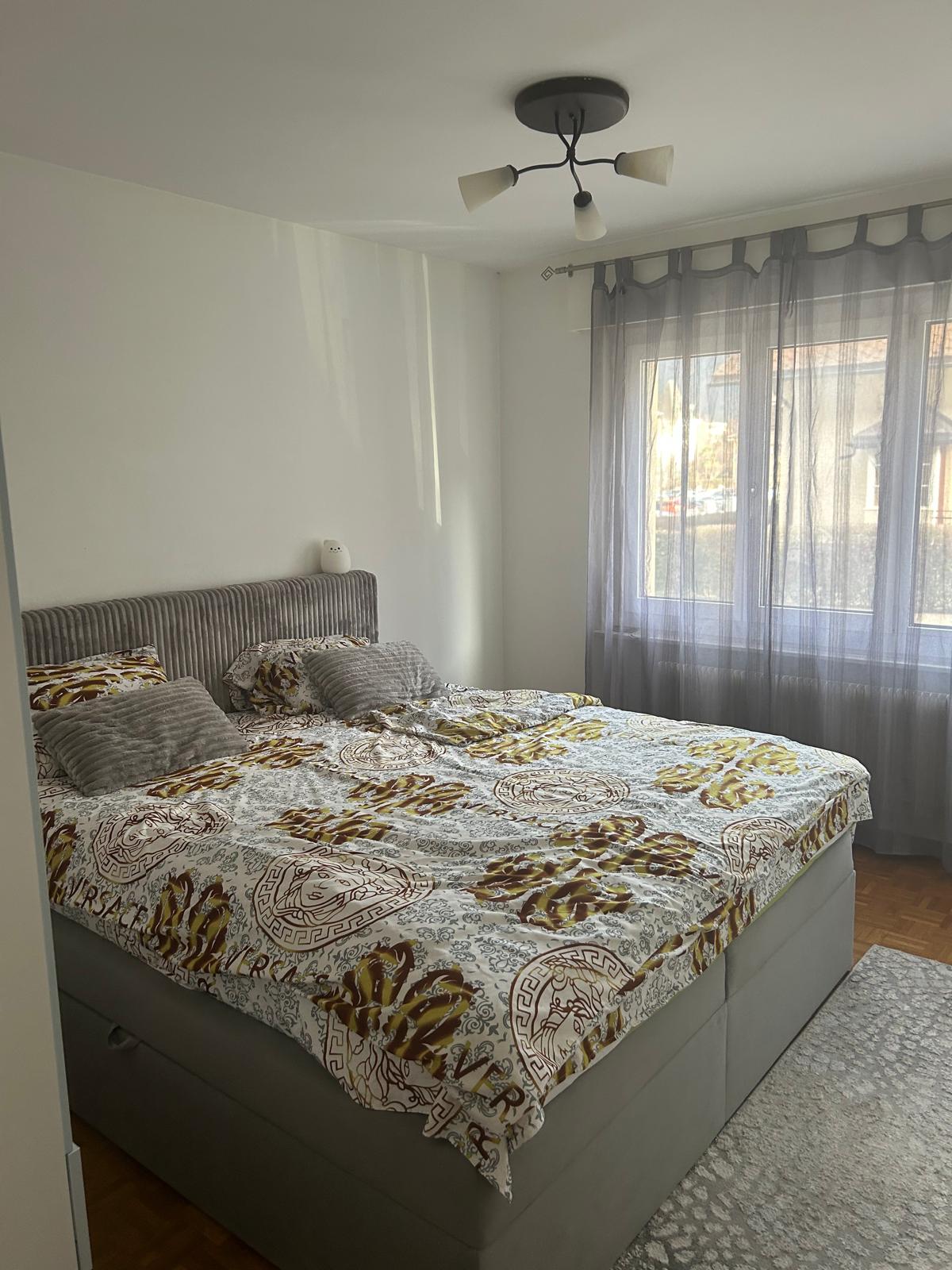 Queen-sized bed with patterned bedding, upholstered headboard, gray walls, white ceiling, large window with curtains, floor lamp, rug