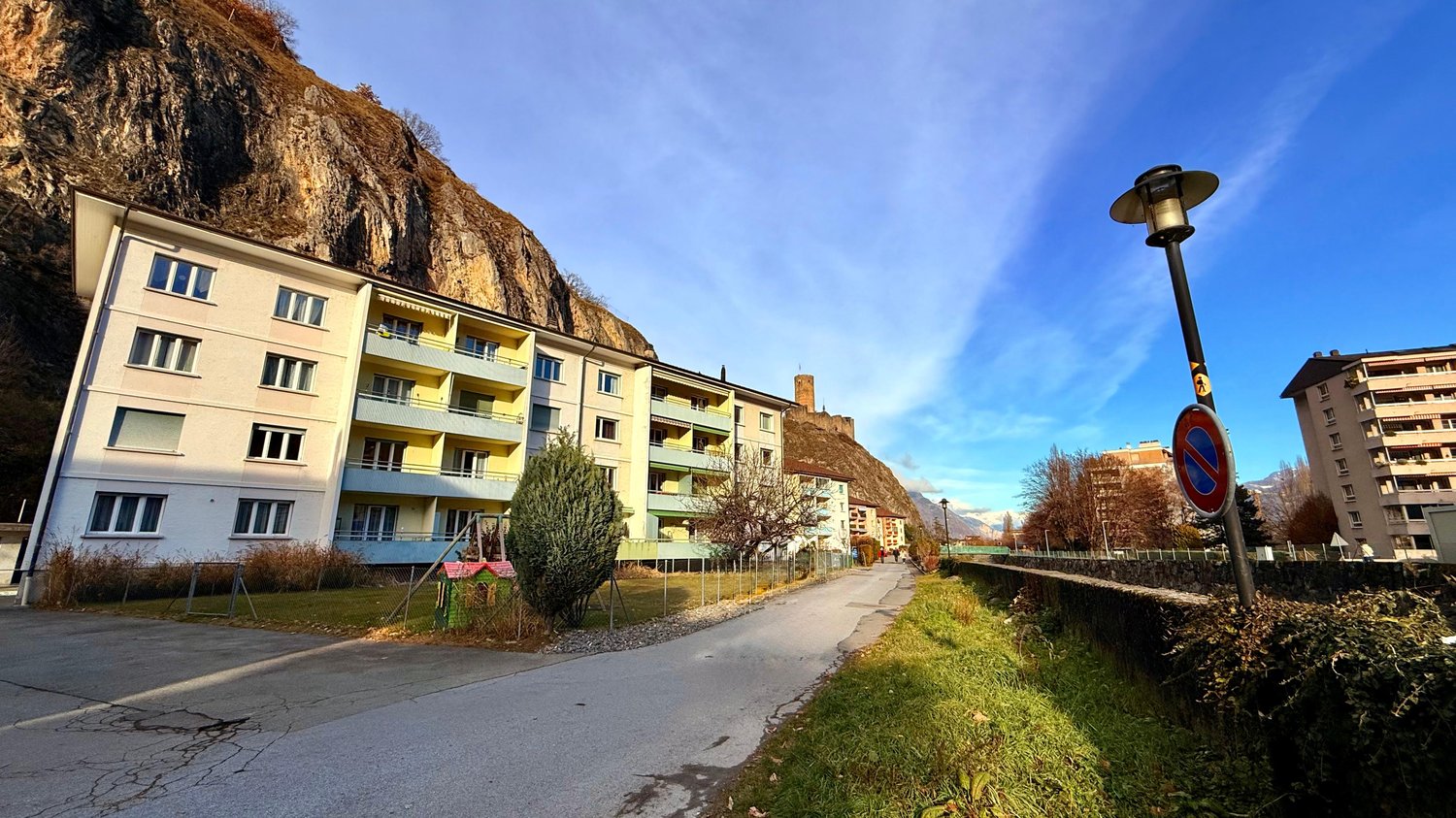 multi-story building, balconies, mountain backdrop, sidewalk, road, parked bicycle, lamp post