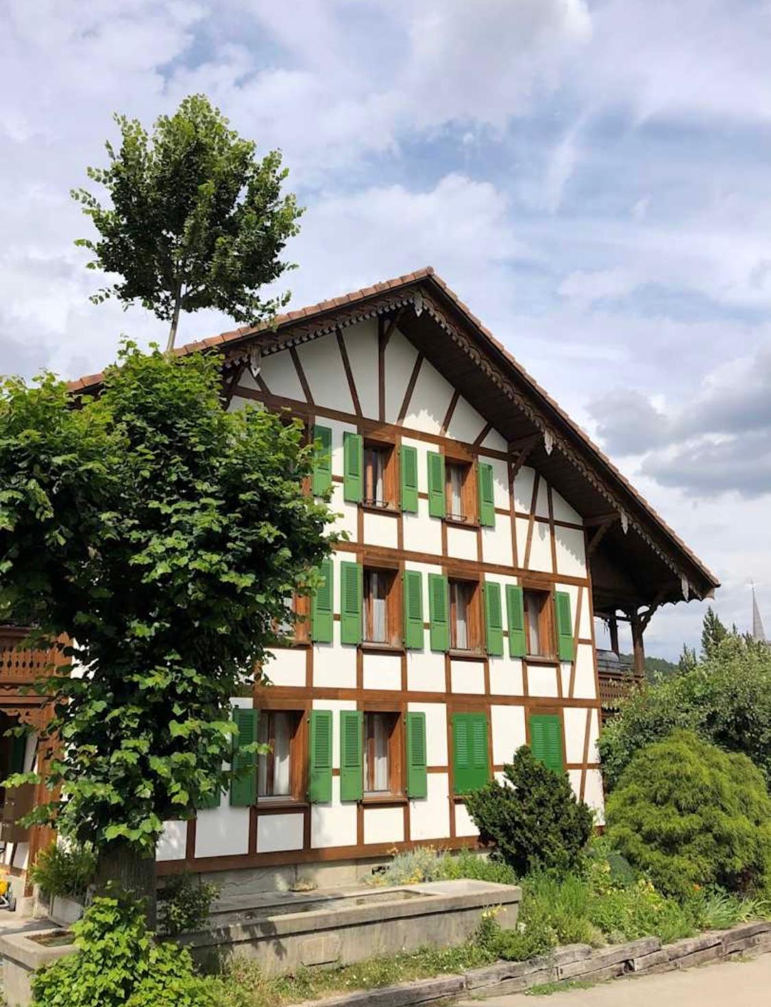 Timber-framed building with green shutters, white paint, tiled roof, large tree in front, small balcony on the left