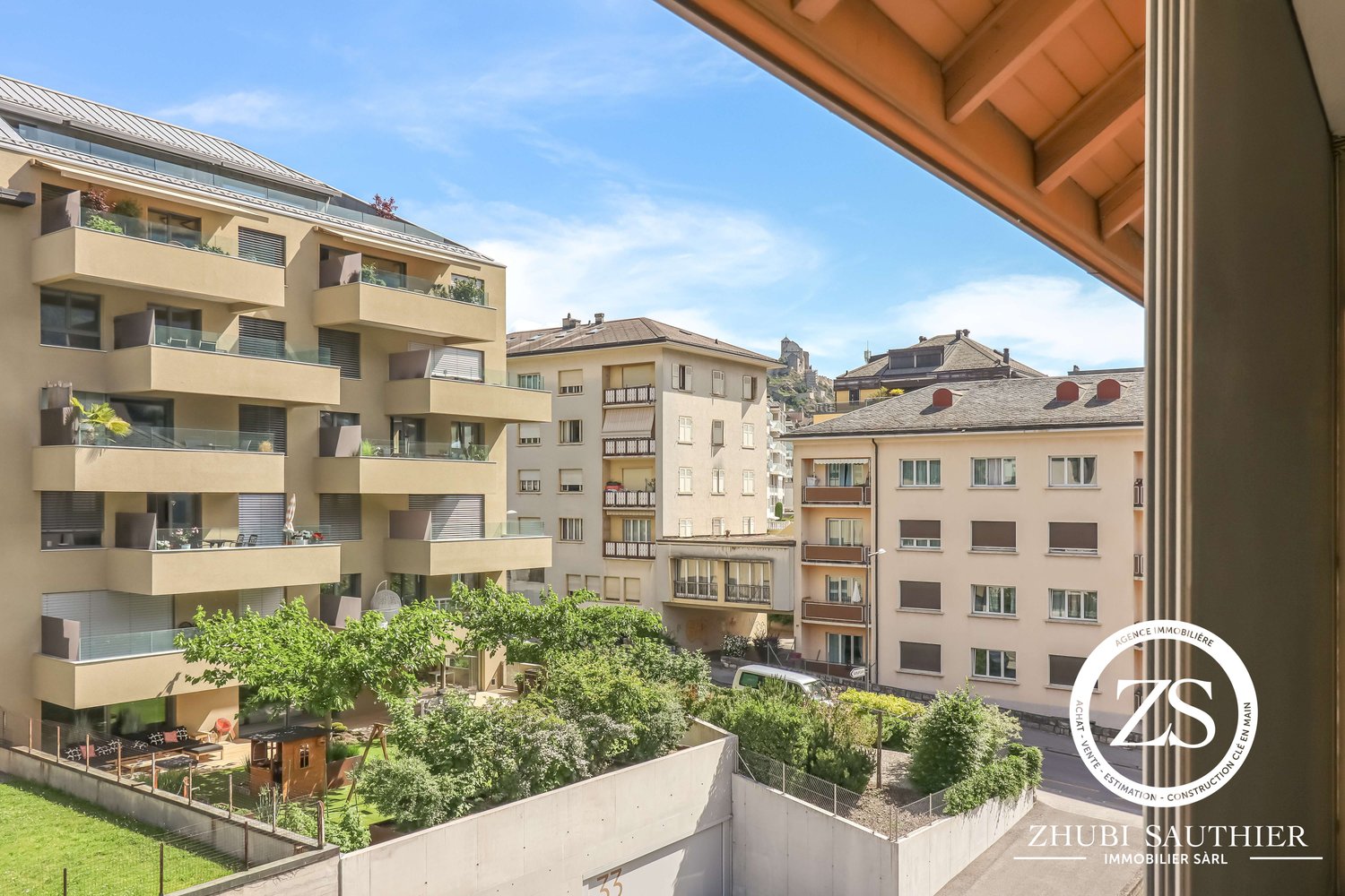 Multi-story apartment building with balconies, surrounded by trees and greenery, with a view of other residential buildings in the background.