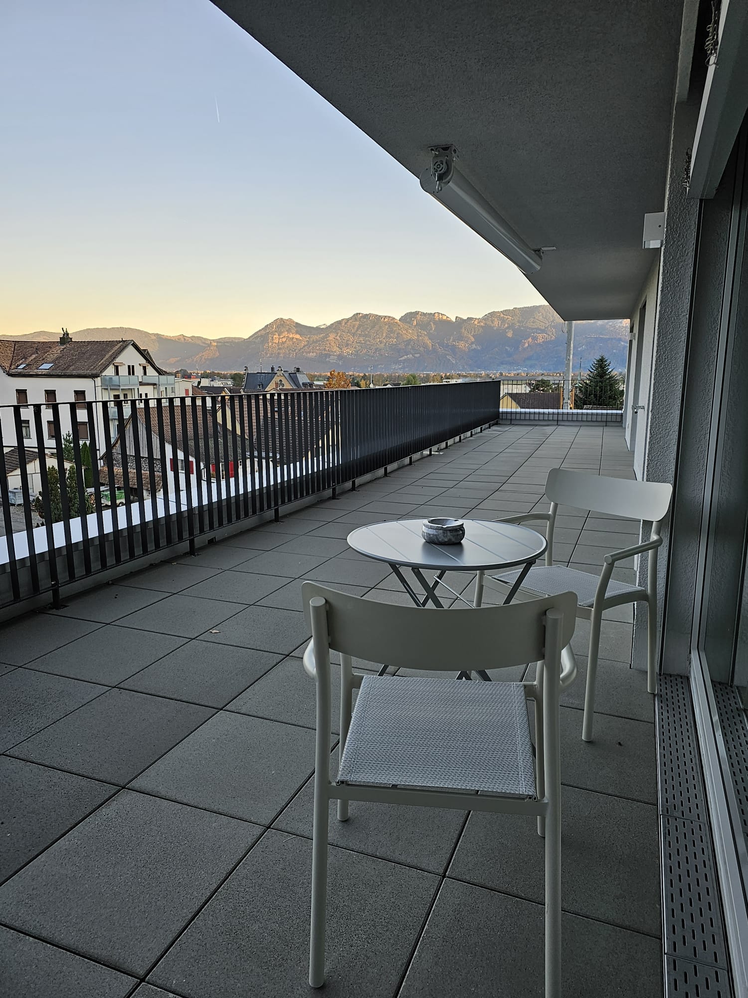 Spacious balcony with a table and chairs, overlooking a scenic mountain landscape in the distance. The balcony has a black railing and the floor is covered with gray tiles.