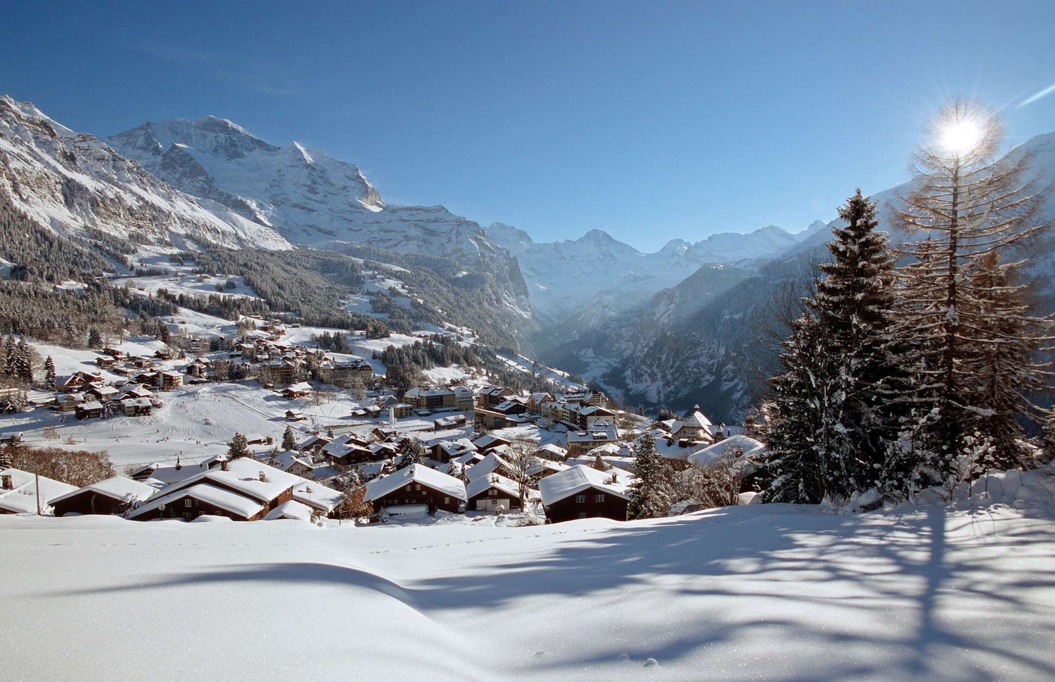 Mountain village with wooden houses, snow-covered roofs, surrounded by snowy mountains, pine trees, and a clear blue sky.