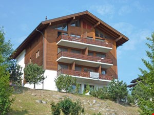 3-story wooden chalet-style apartment building with balconies, situated on a hillside with trees in the foreground
